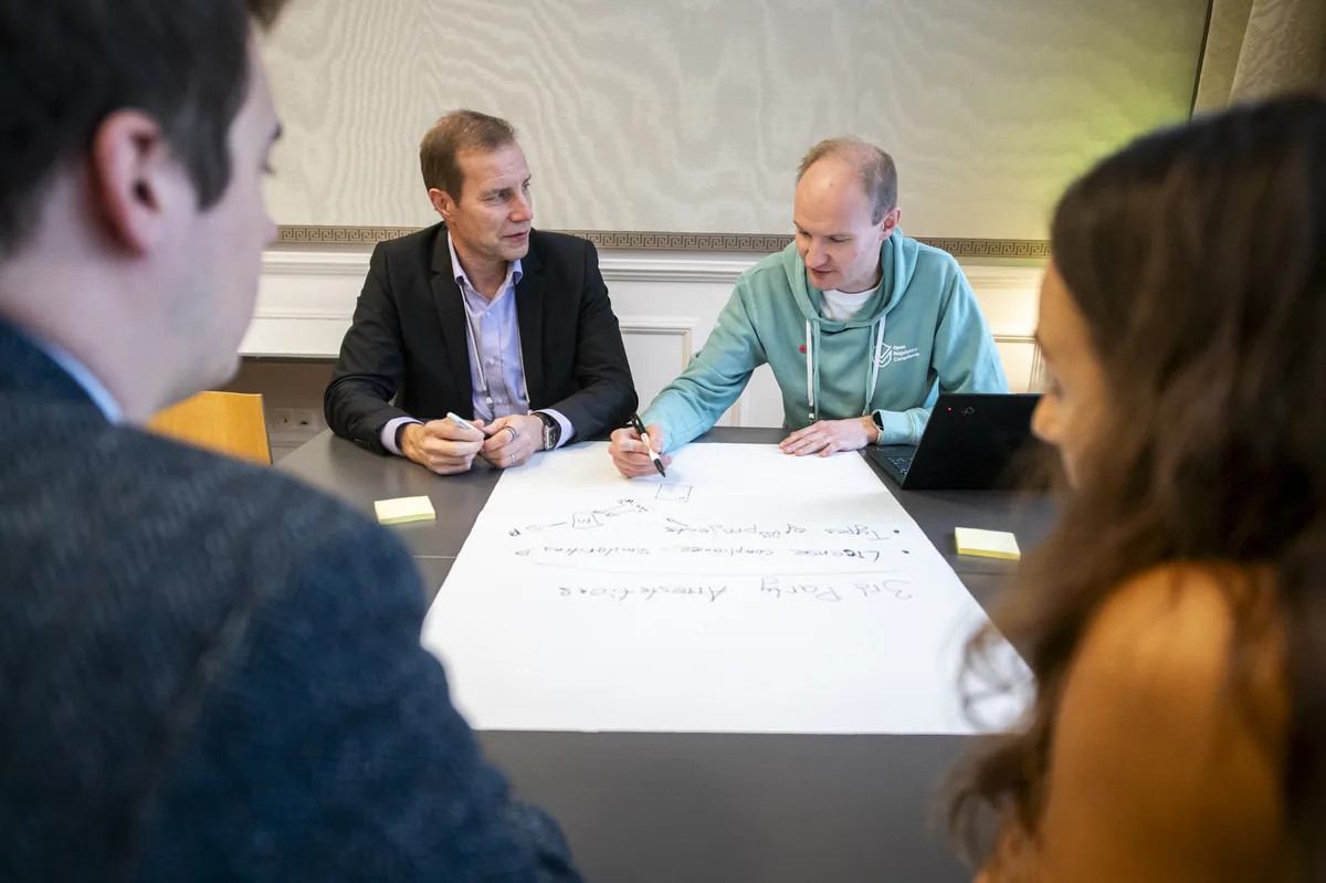 A group of four people collaborating in a room. One person wearing an ORC sweater is writing stuff down on a whiteboard.